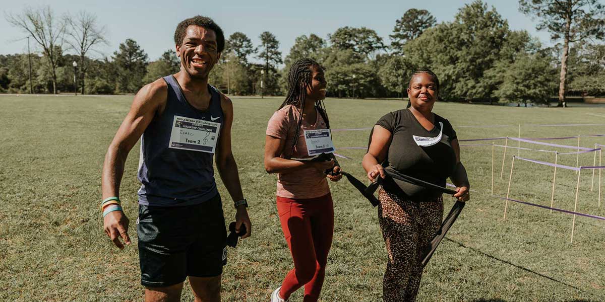 Three MGA students smile at the camera with a runner's ID on their chests labeled Team 2